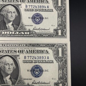 May include: Two United States one dollar silver certificates from series 1957. Each bill features a portrait of George Washington, the text "United States of America", "One Dollar", and a blue treasury seal with the serial numbers B77243894A and B77243893A.