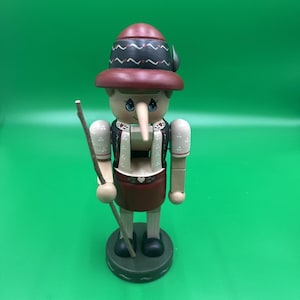 Puede incluir: Una figura de cascanueces de madera con una nariz larga y puntiaguda, que lleva un sombrero rojo oscuro con detalles en blanco y negro, un chaleco negro con corazones blancos y una falda roja oscura. La figura sostiene un bastón de madera.