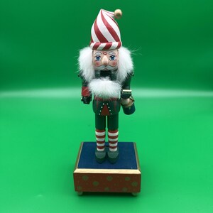 Puede incluir: Una figura de cascanueces de madera con un sombrero de rayas rojas y blancas con una bola dorada en la punta, una chaqueta verde oscuro con detalles rojos y mallas de rayas rojas y blancas. Tiene pelo y barba de piel sintética blanca sobre una base roja.