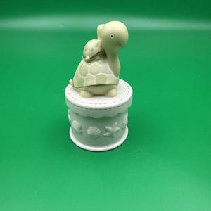 Puede incluir: Una pequeña caja de cerámica con base cilíndrica blanca que presenta patrones en relieve de conchas y estrellas de mar. Una figura de tortuga de color verde pálido con el caparazón bordeado en oro corona la tapa como asa decorativa.