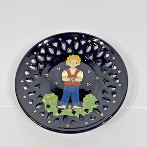 Op de afbeelding: Een donkerblauwe decoratieve schaal met een opengewerkte rand en een geschilderd volkskunstontwerp. De schaal heeft een centraal beeld van een persoon in traditionele kleding, omgeven door groen gebladerte en gele accenten.