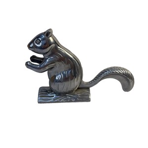 May include: A silver-colored squirrel nutcracker. The squirrel is in a standing position, with its front paws raised. The tail curves upwards. The nutcracker is mounted on a rectangular base with a wood grain texture.
