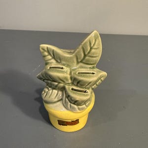 May include: A ceramic coin bank shaped like a plant with large green leaves and a yellow pot. The bank has slots for inserting coins and a small rectangular label on the pot that reads "Miami".