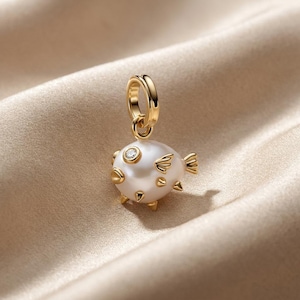 Könnte beinhalten: Ein goldener Anhänger in Form eines Kugelfisches mit einem Körper aus weißer Perle, kleinen goldenen Flossen und einem einzelnen klaren Edelstein als Auge. Der Anhänger besitzt einen runden goldenen Ring zur Befestigung an einer Kette.