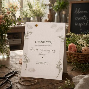 May include: A thank you card for a flower arranging class, with the text "THANK YOU" and "flower arranging class". The card is decorated with leaf illustrations and a gold push pin. A chalkboard with the words "bloom create inspire" is in the background.