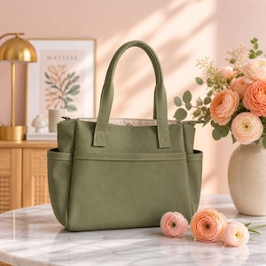 May include: Olive green tote bag with two handles and side pockets. The bag has a zippered top and a structured design. The bag is sitting on a marble table next to a vase of flowers and some loose flowers.