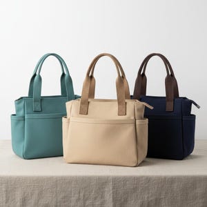 May include: Three canvas tote bags in teal, beige, and navy blue. Each bag has a rectangular shape, two side pockets, and matching handles. The beige and navy bags have a zipper closure. The bags are displayed on a neutral-colored surface.