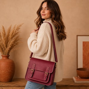 May include: A deep burgundy leather satchel bag featuring a front flap with two buckle closures and an adjustable shoulder strap. The bag has a structured rectangular shape with visible stitching details on the front pockets and flap edges.