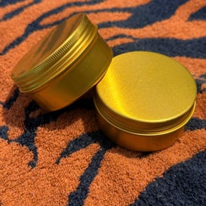 May include: Two round, metallic gold containers with screw-top lids. The containers have a smooth, matte finish and a ribbed edge on the lids for grip. They are positioned side-by-side, with one slightly elevated, showcasing their cylindrical shape.