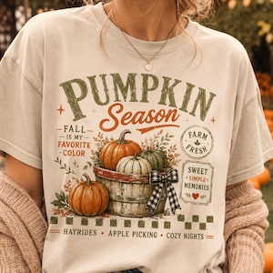 May include: A beige t-shirt featuring a graphic of pumpkins in a wooden basket with a black and white checkered bow. The text reads PUMPKIN Season, FALL IS MY FAVORITE COLOR, FARM FRESH, SWEET SIMPLE MEMORIES, HAYRIDES, APPLE PICKING, COZY NIGHTS.