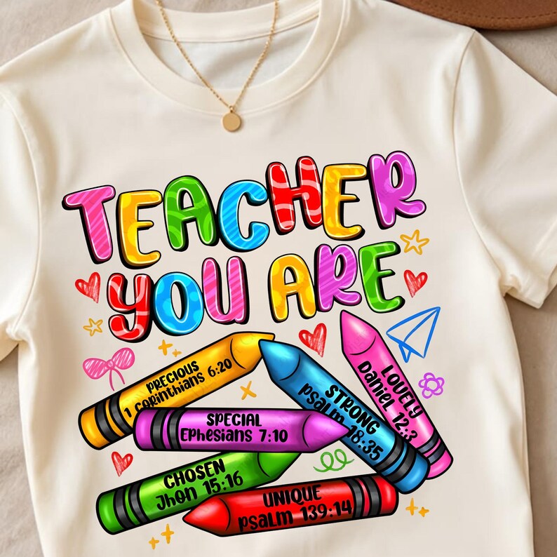 May include: A cream-colored t-shirt featuring the colorful text TEACHER YOU ARE above five crayons in yellow, blue, pink, green, and red. Each crayon displays a word and a Bible verse, including Precious 1 Corinthians 6:20, Strong Psalm 18:35, Lovely Daniel 12:3, Special Ephesians 7:10, Chosen John 15:16, and Unique Psalm 139:14.