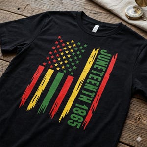 Peut inclure: T-shirt noir avec un motif de drapeau stylisé aux couleurs rouge, jaune, vert et or. Le motif du drapeau comprend des étoiles et des rayures, avec les mots "Juneteenth 1865" imprimés en diagonale en vert et rouge.