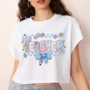 May include: A white short-sleeved t-shirt featuring a graphic print with the word TEACHER in bubble letters. The design includes a light blue checkered background, white daisies, small stars, and a large light blue bow tied around a pencil.