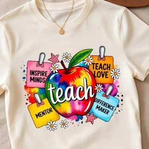 May include: A cream-colored t-shirt featuring a central graphic of a rainbow-colored apple with the word teach written in white script. Surrounding the apple are four colorful sticky notes labeled inspire minds, teach love, mentor, and difference maker.