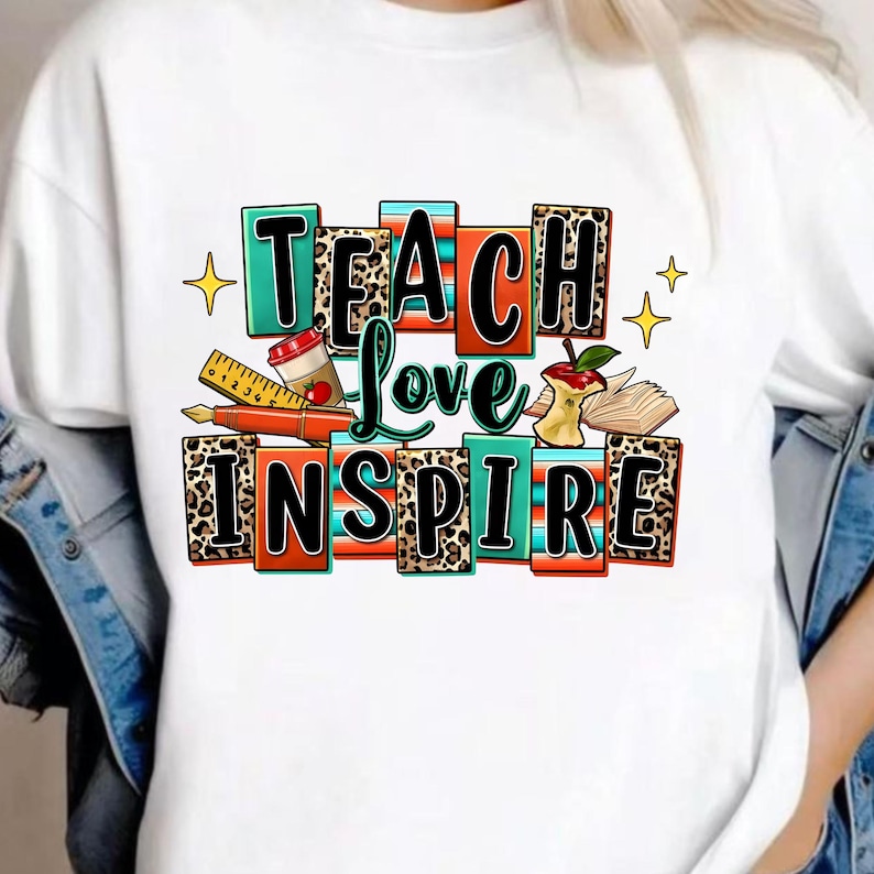 May include: A white t-shirt featuring a colorful graphic with the text TEACH LOVE INSPIRE. The design includes school-themed illustrations like a ruler, a fountain pen, a coffee cup, an apple core, and an open book, set against leopard print.