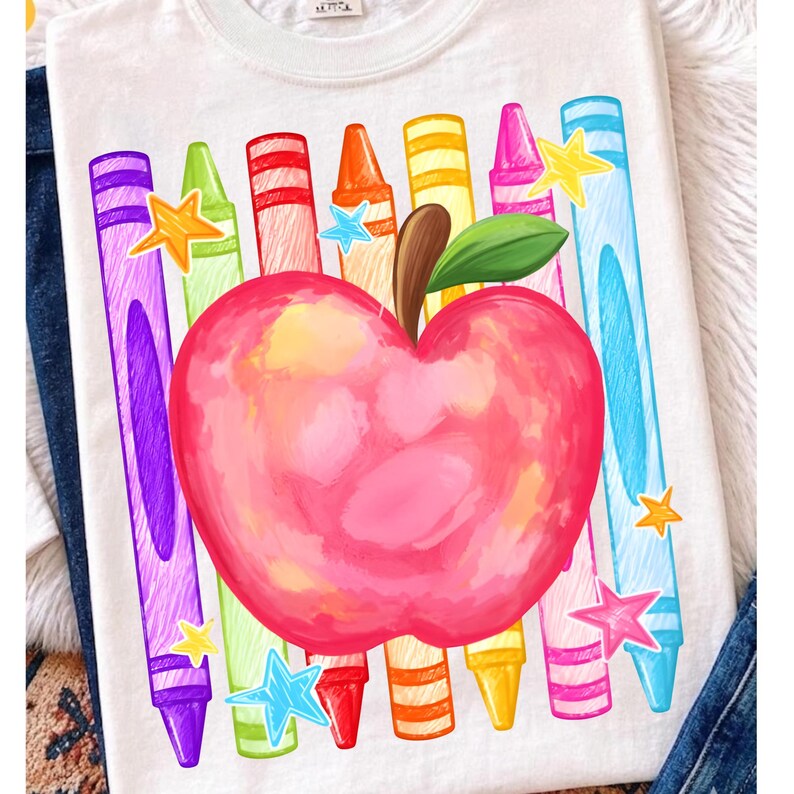 May include: A white t-shirt featuring a large, watercolor-style red apple with a green leaf in the center. Surrounding the apple are vertical, colorful crayon illustrations in purple, green, red, orange, yellow, pink, and blue, accented by small stars.