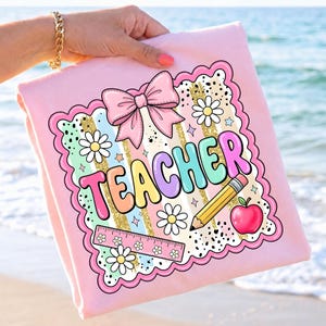 May include: A light pink t-shirt featuring a graphic design with the word TEACHER in colorful bubble letters. The design includes a large pink bow, white daisies, a yellow pencil, a red apple, and a ruler, all framed by a scalloped border.