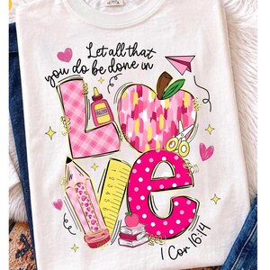 May include: A white t-shirt featuring the text Let all that you do be done in love 1 Cor 16:14. The graphic includes a pink plaid letter L, a pink brushstroke apple, a yellow ruler, a pink polka dot letter E, a pencil, glue, and scissors.