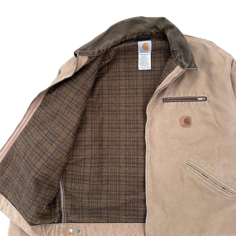 Peut inclure: Veste Carhartt beige avec un col en velours c&ocirc;tel&eacute; marron et une doublure &agrave; carreaux marron. La veste a une fermeture &eacute;clair, une poche poitrine et un petit &eacute;cusson logo Carhartt. La veste est ouverte, r&eacute;v&eacute;lant la doublure int&eacute;rieure.