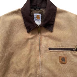 Peut inclure: Veste Carhartt beige avec un col en velours c&ocirc;tel&eacute; marron et une fermeture &eacute;clair int&eacute;grale. La veste pr&eacute;sente une poche marron et un &eacute;cusson logo Carhartt. La doublure int&eacute;rieure est en tissu &agrave; motifs. Style workwear.