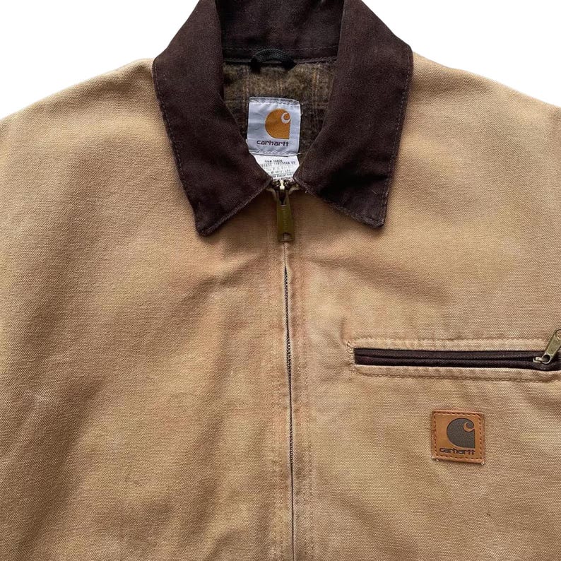 Peut inclure: Veste Carhartt beige avec un col en velours c&ocirc;tel&eacute; marron et une fermeture &eacute;clair int&eacute;grale. La veste pr&eacute;sente une poche marron et un &eacute;cusson logo Carhartt. La doublure int&eacute;rieure est en tissu &agrave; motifs. Style workwear.