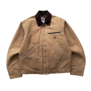 May include: A tan work jacket with a brown corduroy collar, a zip closure, and multiple pockets. The jacket features the Carhartt logo on the chest and a small zip pocket on the chest. The jacket is made of a durable, heavy-duty material.