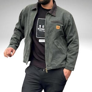 May include: A dark green Carhartt jacket with a brown corduroy collar and a small square logo patch. The jacket is zipped up and has a chest pocket. The person is wearing a black t-shirt with the word "BURBERRY" in a white rectangle.