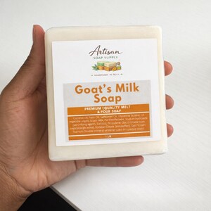 Op de afbeelding: Een rechthoekige zeepblok van geitenmelk, vastgehouden in een hand. De zeep heeft een wit etiket met de woorden "Artisan Soap Supply" en "Goat's Milk Soap". Het etiket vermeldt ook ingredi&euml;nten en het gewicht, 454 gram.
