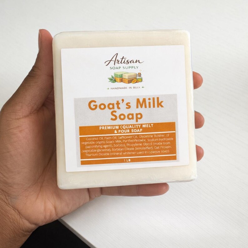 Op de afbeelding: Een rechthoekige zeepblok van geitenmelk, vastgehouden in een hand. De zeep heeft een wit etiket met de woorden "Artisan Soap Supply" en "Goat's Milk Soap". Het etiket vermeldt ook ingredi&euml;nten en het gewicht, 454 gram.
