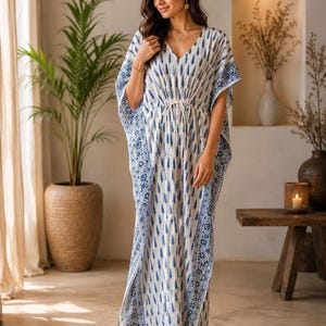 May include: A floor-length kaftan dress featuring a white base with a repeating blue tree pattern. The garment has a V-neckline, wide short sleeves, and a drawstring waist with white ties. The edges are finished with a blue and white floral border.