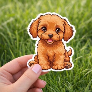 Peut inclure: Autocollant représentant un chiot brun de dessin animé, avec de grands yeux et une expression joyeuse. Le chiot est assis, la langue visible. L'autocollant est tenu dans une main, sur fond d'herbe verte.