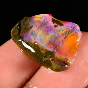 Könnte beinhalten: Ein lebendiger, roher Opal-Edelstein mit einem Farbspektrum aus Orange, Rot, Grün und Lila. Der Opal hat eine raue, natürliche Textur mit einer dunkelgrünen Außenschicht. Der Edelstein wird in der Hand einer Person vor schwarzem Hintergrund gehalten.