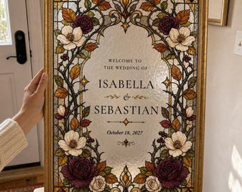 May include: A rectangular wedding sign in a gold frame featuring a stained glass style border with white flowers, dark red roses, and autumn leaves. The centre text reads: WELCOME TO THE WEDDING OF ISABELLA & SEBASTIAN, October 18, 2027.