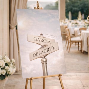 Könnte beinhalten: Ein Hochzeits-Willkommensschild auf einer goldenen Staffelei. Das Schild zeigt ein Straßenschild-Design mit den Namen GARCIA und DELACRUZ, mit dem Text "Where it all began" und dem Datum. Ein Blumenarrangement befindet sich in einer Steinvase.