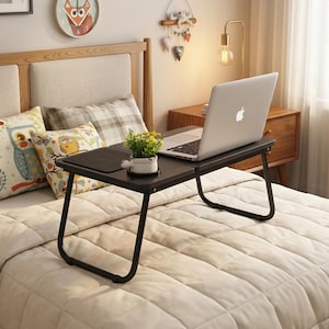 May include: A black folding laptop desk with a laptop on top, placed on a bed. The desk features a mouse section and a small white plant pot. The bed has pillows and a decorative headboard.