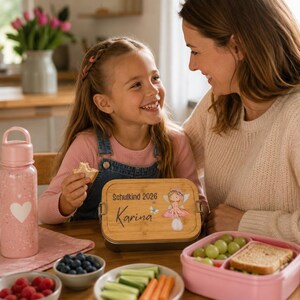 Puede incluir: Una fiambrera rectangular con tapa de bamb&uacute; que presenta el texto "Schulkind 2026" y "Karina" junto a una ilustraci&oacute;n de hada, acompa&ntilde;ada de una botella de agua rosa claro decorada con un coraz&oacute;n blanco y un recipiente rosa dividido.