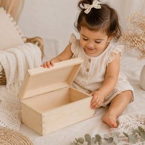 May include: A light-colored wooden box with a hinged lid is open, revealing an empty interior. The box is rectangular and appears to be made of unfinished wood. The box is sitting on a white surface.