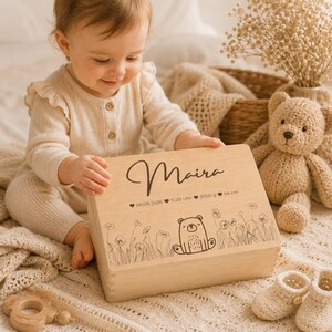 May include: A light-colored wooden box with the name "Maira" in cursive. The box is decorated with a bear illustration and floral designs. The box also has the date, time, weight, and height of the baby. A baby is holding the box.