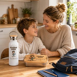 May include: A white metal water bottle with a bamboo lid and a matching rectangular metal lunch box with a bamboo lid. Both items feature a printed illustration of a child with a globe, the text Schulkind 2026, and the name Adrian in script.