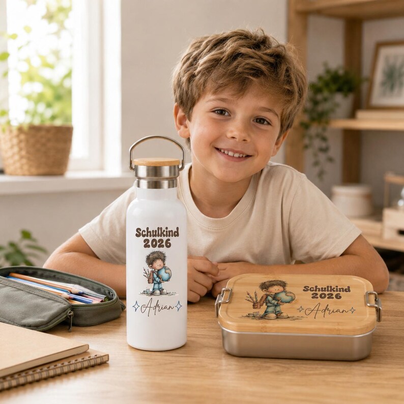 May include: A white metal water bottle with a bamboo lid and a rectangular metal lunch box with a bamboo lid. Both items feature a cartoon illustration of a child, the text "Schulkind 2026", and the name "Adrian" printed in a dark brown font.