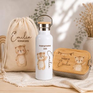 May include: A beige drawstring bag, a white metal water bottle with a silver handle and bamboo lid, and a rectangular metal lunch box with a bamboo lid. Each item features a brown bear illustration and the name Emilia printed in black text.