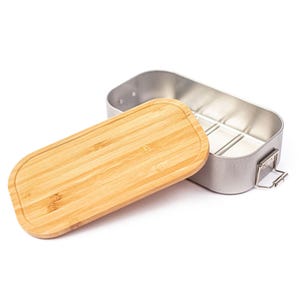 Puede incluir: Una fiambrera rectangular de metal con una tapa de bamb&uacute; lisa de color marr&oacute;n claro. El recipiente presenta un acabado plateado met&aacute;lico, esquinas redondeadas y un peque&ntilde;o mecanismo de cierre de alambre en el lateral para fijar la tapa.