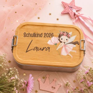 Puede incluir: Una fiambrera rectangular con tapa de bamb&uacute; de color marr&oacute;n claro que presenta el texto impreso Schulkind 2026 y el nombre Laura, junto a una ilustraci&oacute;n en acuarela de un hada con alas rosas y una flor, asegurada por dos cierres met&aacute;licos.