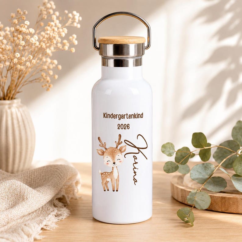 Könnte beinhalten: Eine weiße Metalltrinkflasche mit silbernem Rand und einem Holzdeckel mit Metallbügel. Die Flasche ist mit einer braunen Illustration eines kleinen Rehs, dem Text "Kindergartenkind 2026" und dem Namen "Karina" in Schreibschrift bedruckt.