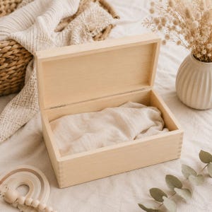 May include: An open, light-colored wooden box with a hinged lid. Inside the box is a folded, beige fabric. The box is rectangular with visible dovetail joints. A woven basket and a vase with dried flowers are in the background.