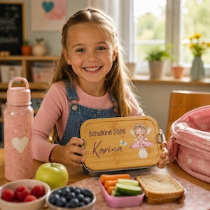 Puede incluir: Una fiambrera rectangular con tapa de bamb&uacute; de color marr&oacute;n claro que presenta el texto impreso Schulkind 2026 y el nombre Karina, junto con un gr&aacute;fico de un hada sentada sobre una seta. La fiambrera se acompa&ntilde;a de una botella de agua rosa.