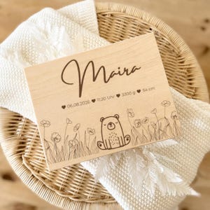 May include: A light-colored wooden plaque with the name "Maira" in cursive script. The plaque includes the date, time, weight (3300 g), and height (54 cm) of a baby, along with a bear and floral design.