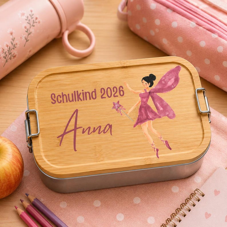 Puede incluir: Una fiambrera rectangular con tapa de bamb&uacute; claro que presenta una ilustraci&oacute;n de un hada con vestido morado y una varita de estrella. El texto "Schulkind 2026" y el nombre "Anna" est&aacute;n impresos en morado. La base es de metal plateado.