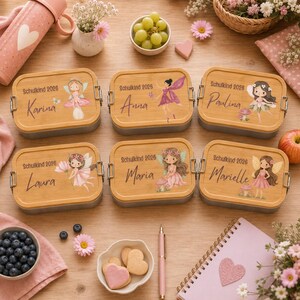 Puede incluir: Seis fiambreras rectangulares con tapas de bamb&uacute;, cada una con una ilustraci&oacute;n de hadas, el texto "Schulkind 2026" y un nombre. Las cajas est&aacute;n dispuestas sobre una superficie de madera junto a un cuenco de uvas, galletas y un cuaderno.