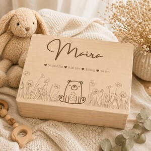 May include: A light-colored wooden box with the name "Maira" engraved on the lid. The box features a bear illustration and floral designs. The lid also includes the date 06.08.2026, time 11:20 Uhr, weight 3300 g, and height 54 cm.